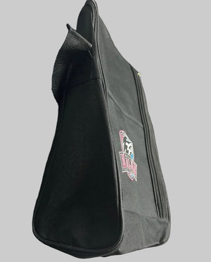 A black canvas shoe bag with a small pink and white logo patch, resting on a gray workbench in a storage or warehouse setting with cardboard boxes in the background.